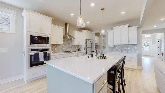 kitchen with wall chimney exhaust hood, a center island with sink, pendant lighting, stainless steel appliances, and white cabinets