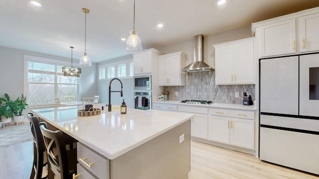 kitchen featuring refrigerator, an island with sink, white cabinets, gas stovetop, and wall chimney exhaust hood