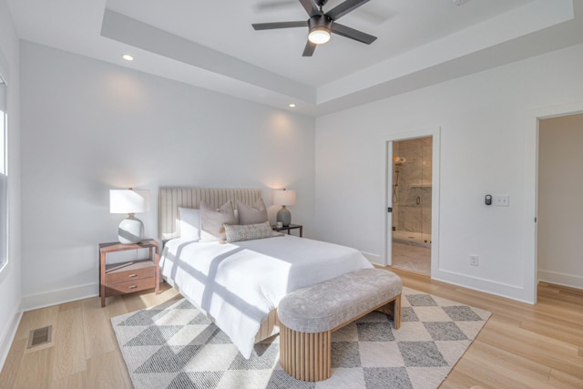 bedroom featuring ceiling fan, ensuite bathroom, a raised ceiling, and light wood-type flooring