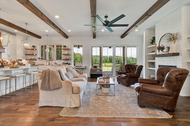 living room with sink, beam ceiling, dark hardwood / wood-style flooring, a brick fireplace, and ceiling fan with notable chandelier