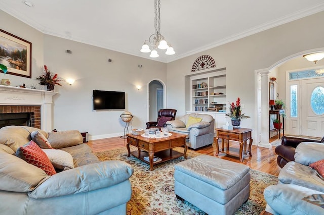 living room with crown molding, an inviting chandelier, a brick fireplace, built in shelves, and light wood-type flooring