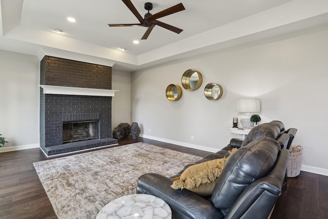 living room with dark wood-type flooring, ceiling fan, a raised ceiling, and a brick fireplace