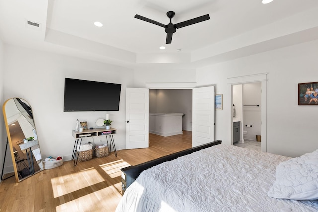 bedroom with a raised ceiling, ensuite bathroom, ceiling fan, and light wood-type flooring