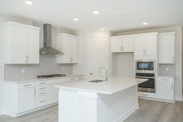 kitchen with appliances with stainless steel finishes, an island with sink, sink, white cabinets, and wall chimney exhaust hood