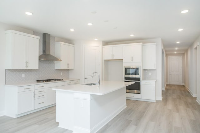 kitchen featuring an island with sink, sink, white cabinets, stainless steel appliances, and wall chimney range hood