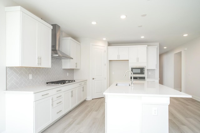 kitchen with wall chimney exhaust hood, sink, gas stovetop, white cabinetry, and an island with sink