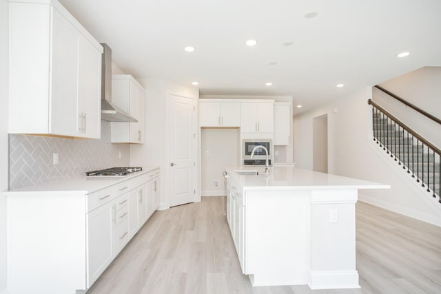 kitchen with sink, light hardwood / wood-style flooring, an island with sink, white cabinets, and wall chimney exhaust hood