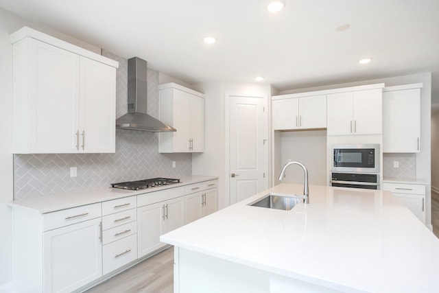 kitchen featuring sink, appliances with stainless steel finishes, an island with sink, wall chimney range hood, and white cabinets