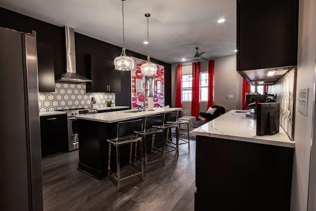 kitchen with decorative light fixtures, wall chimney range hood, stainless steel appliances, a kitchen island with sink, and backsplash