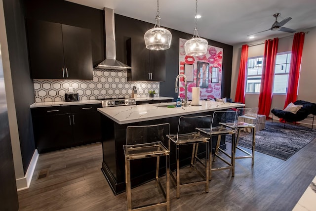 kitchen featuring wall chimney exhaust hood, sink, decorative light fixtures, a center island with sink, and backsplash