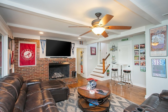 living room featuring ceiling fan, hardwood / wood-style floors, beam ceiling, a fireplace, and brick wall