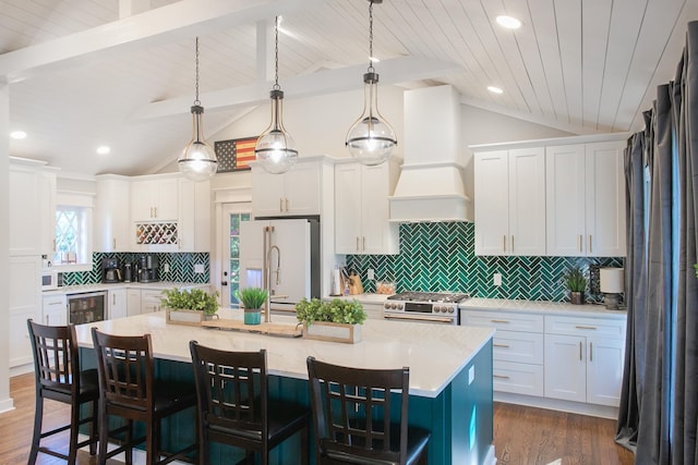 kitchen with an island with sink, white cabinets, hanging light fixtures, high end fridge, and custom range hood