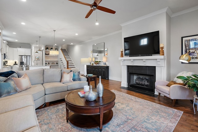 living room featuring sink, a tiled fireplace, ceiling fan, crown molding, and dark wood-type flooring