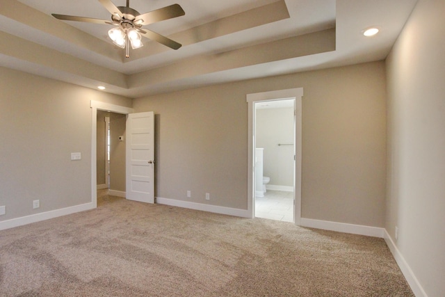 unfurnished bedroom with a raised ceiling, ensuite bathroom, light colored carpet, and ceiling fan