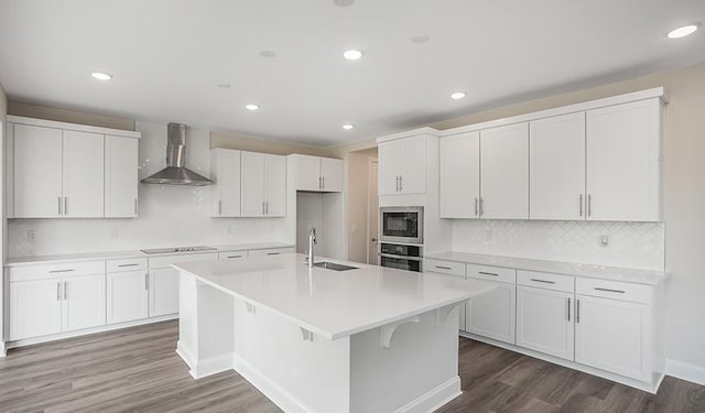 kitchen featuring built in microwave, white cabinetry, oven, a kitchen island with sink, and wall chimney range hood