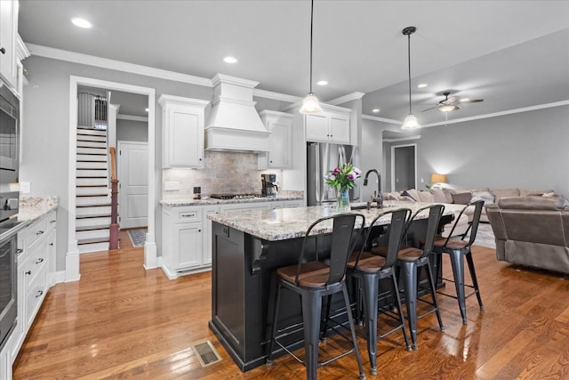 kitchen featuring a kitchen island with sink, stainless steel appliances, custom range hood, and white cabinets