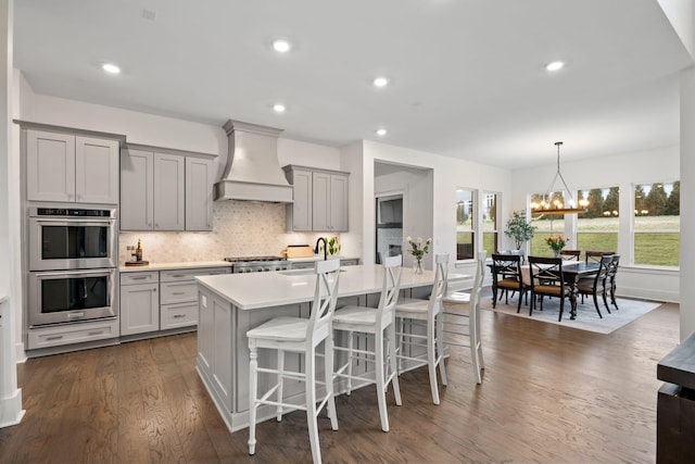 kitchen with premium range hood, a kitchen breakfast bar, gray cabinets, stainless steel double oven, and a kitchen island with sink