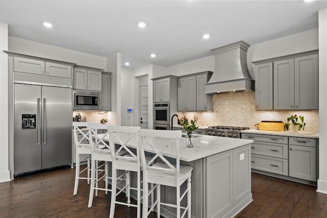 kitchen with built in appliances, gray cabinetry, custom range hood, and an island with sink