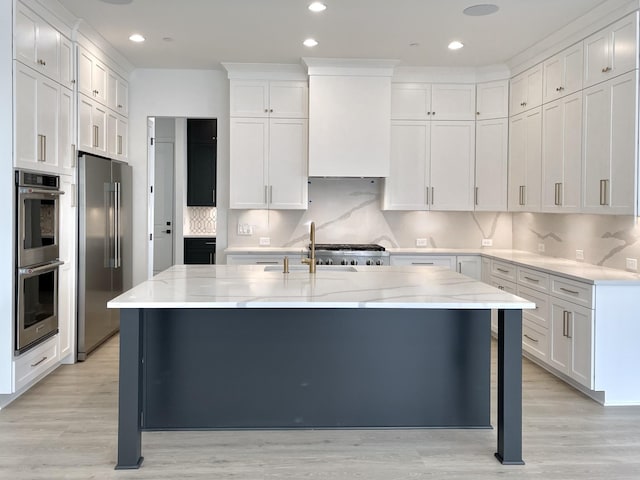 kitchen with appliances with stainless steel finishes, white cabinetry, a center island with sink, decorative backsplash, and light wood-type flooring