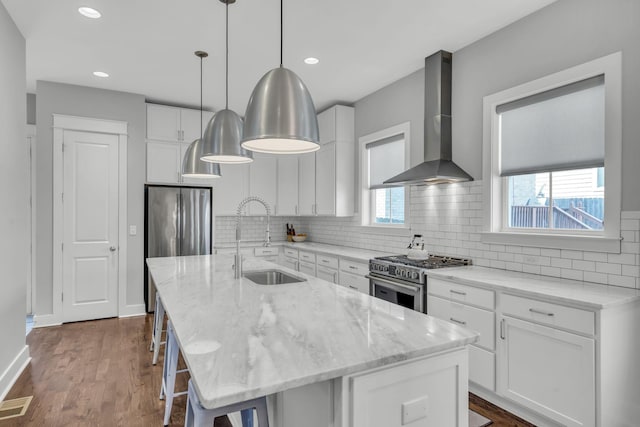 kitchen with wall chimney range hood, decorative light fixtures, stainless steel appliances, and an island with sink