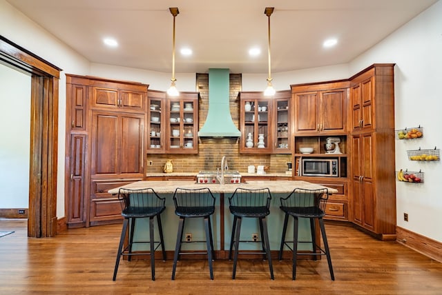 kitchen with dark hardwood / wood-style floors, pendant lighting, stainless steel microwave, a kitchen island with sink, and custom range hood