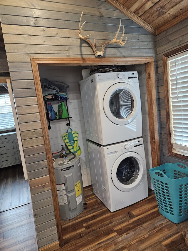 washroom featuring water heater, stacked washer / drying machine, dark wood-type flooring, and wooden walls