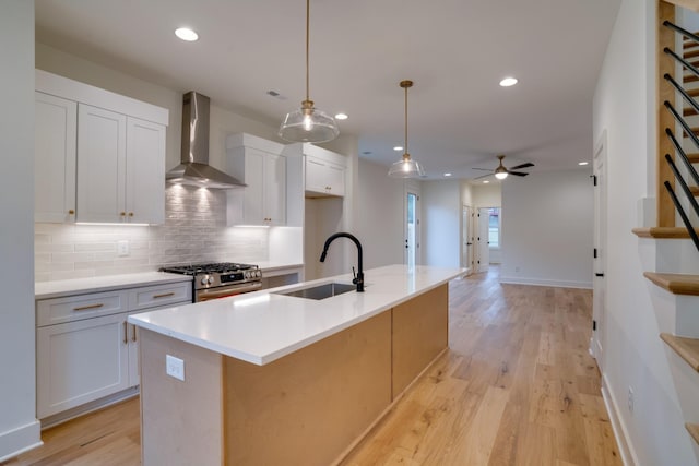 kitchen with an island with sink, sink, white cabinets, stainless steel gas range oven, and wall chimney range hood