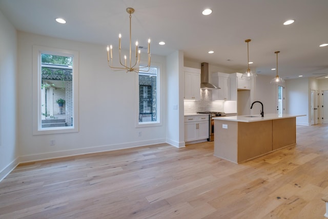 kitchen featuring wall chimney range hood, a kitchen island with sink, hanging light fixtures, stainless steel gas range oven, and white cabinets