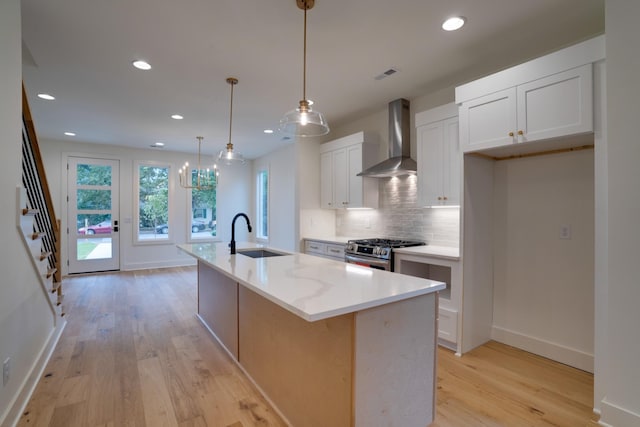 kitchen featuring wall chimney exhaust hood, sink, stainless steel range with gas stovetop, an island with sink, and white cabinets