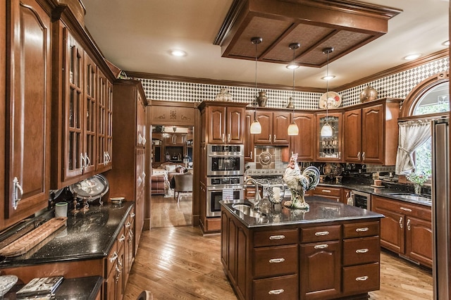 kitchen with hanging light fixtures, an island with sink, dark stone counters, stainless steel double oven, and light wood-type flooring