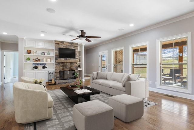 living room with built in shelves, a stone fireplace, crown molding, ceiling fan, and light hardwood / wood-style floors