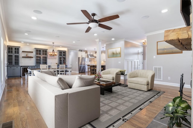 living room with ceiling fan with notable chandelier, decorative columns, beverage cooler, crown molding, and light wood-type flooring