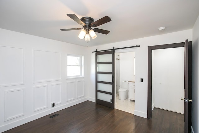 unfurnished bedroom featuring ensuite bath, dark wood-type flooring, a barn door, and ceiling fan