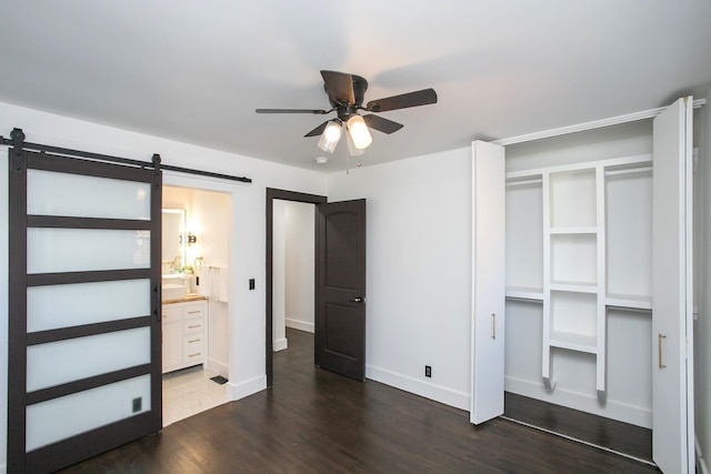 unfurnished bedroom featuring ceiling fan, connected bathroom, a barn door, and dark hardwood / wood-style flooring