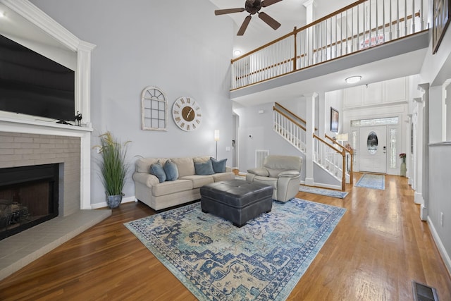 living room with a towering ceiling, decorative columns, wood-type flooring, ceiling fan, and a brick fireplace