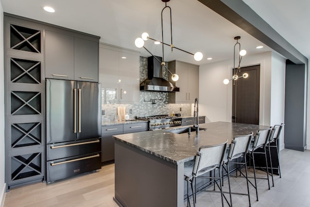 kitchen with sink, hanging light fixtures, high quality appliances, dark stone counters, and wall chimney exhaust hood