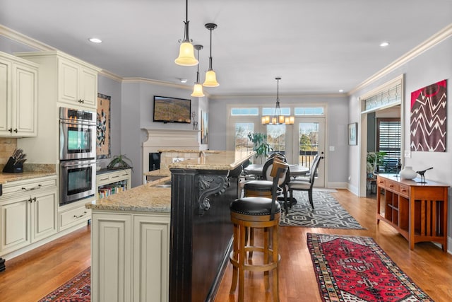 kitchen with a kitchen island with sink, double oven, light stone counters, cream cabinets, and decorative light fixtures