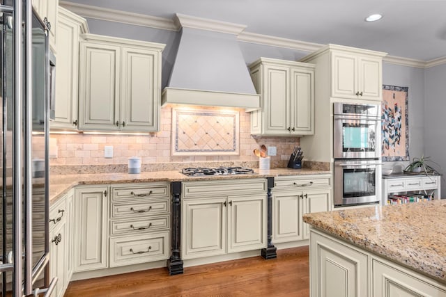 kitchen featuring crown molding, appliances with stainless steel finishes, custom range hood, light stone countertops, and cream cabinets