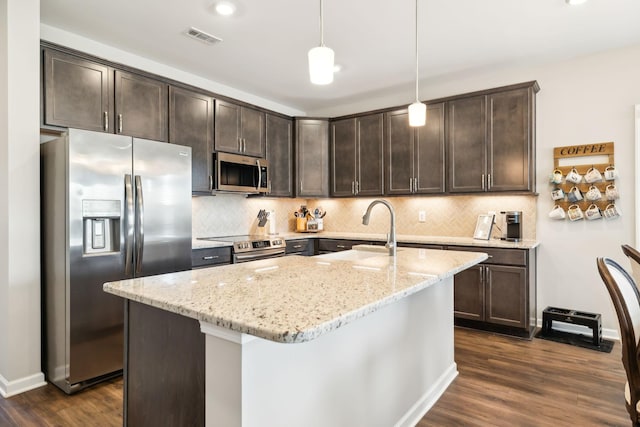 kitchen with appliances with stainless steel finishes, decorative light fixtures, an island with sink, light stone countertops, and dark brown cabinets