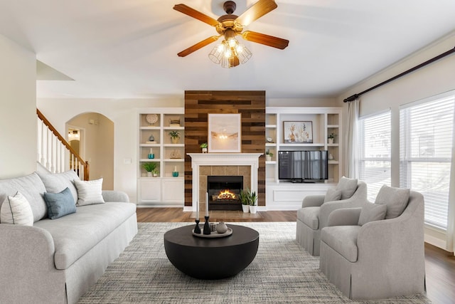 living room with dark wood-type flooring, ceiling fan, a tiled fireplace, and built in shelves