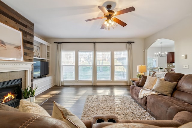 living room featuring a tiled fireplace, built in shelves, dark wood-type flooring, and ceiling fan with notable chandelier