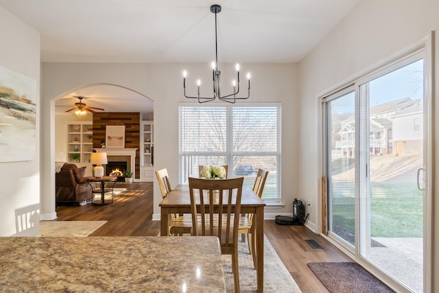 dining space with a tiled fireplace, built in shelves, wood-type flooring, and ceiling fan with notable chandelier