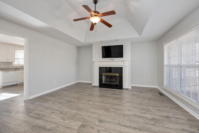 unfurnished living room featuring ceiling fan, a tray ceiling, a tiled fireplace, and light hardwood / wood-style floors