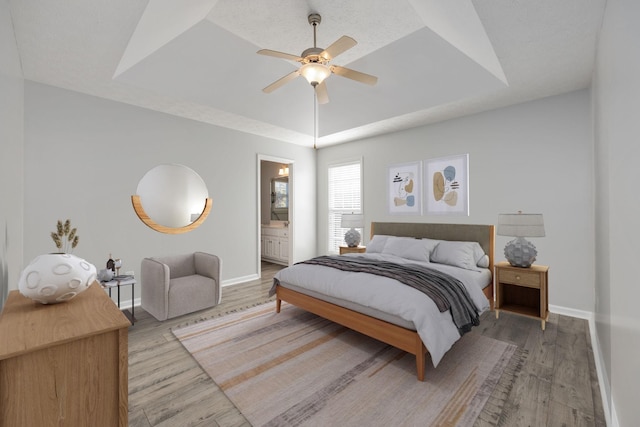 bedroom with ensuite bath, light wood-type flooring, ceiling fan, and a tray ceiling