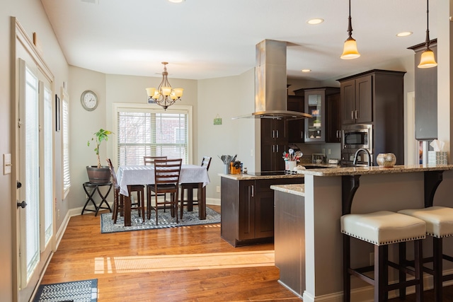 kitchen with hanging light fixtures, stainless steel microwave, light stone counters, dark brown cabinetry, and island exhaust hood