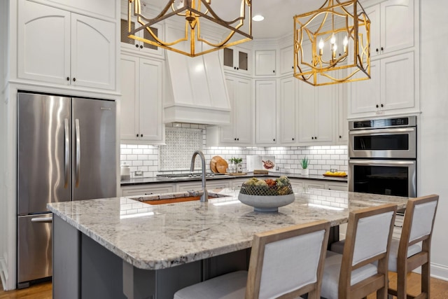 kitchen with appliances with stainless steel finishes, a kitchen island with sink, hanging light fixtures, an inviting chandelier, and custom range hood