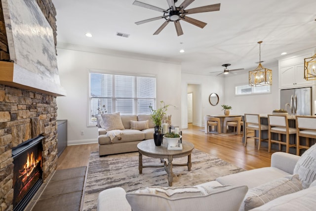 living room with crown molding, a stone fireplace, ceiling fan with notable chandelier, and light hardwood / wood-style flooring