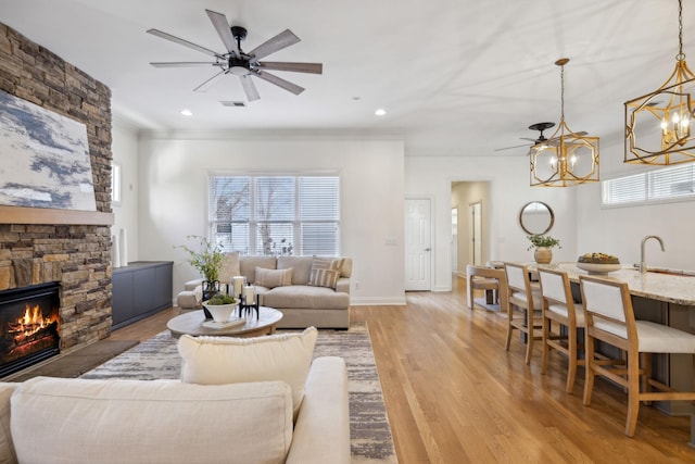 living room with ceiling fan with notable chandelier, a fireplace, sink, ornamental molding, and light wood-type flooring