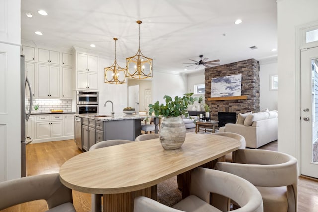 dining room with a stone fireplace, sink, crown molding, light wood-type flooring, and ceiling fan with notable chandelier