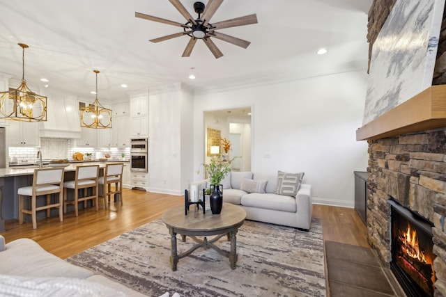 living room featuring sink, light wood-type flooring, ornamental molding, a fireplace, and ceiling fan with notable chandelier